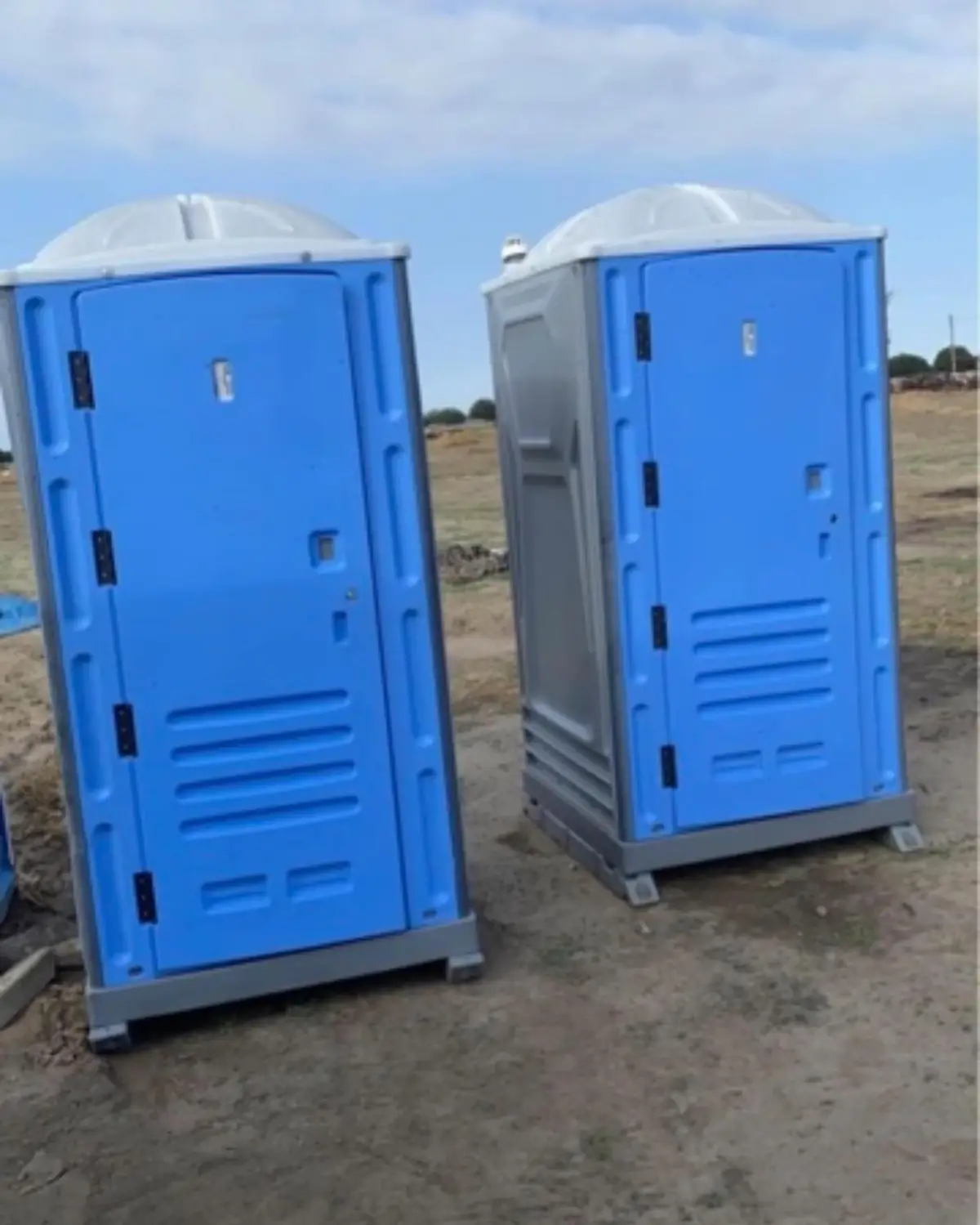 Two clean blue standard porta potties with translucent roofs standing on a dirt jobsite